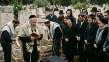 Group of mourners attending a Jewish burial ceremony with rabbi reading from a prayer book near a coffin.